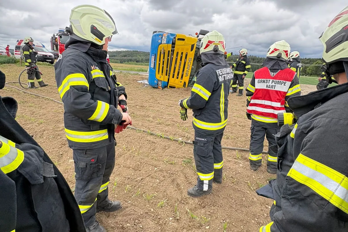 Ein umgestürzter blauer und gelber Lastkraftwagen liegt auf einem Feld, umgeben von Feuerwehrleuten in Schutzkleidung. Der Himmel ist bewölkt, und im Hintergrund sind weitere Einsatzfahrzeuge zu sehen.