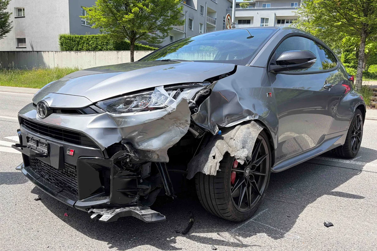 Ein silbernes Auto mit erheblichen Schäden an der Front steht auf einer Straße. Im Hintergrund sind Wohngebäude sichtbar.