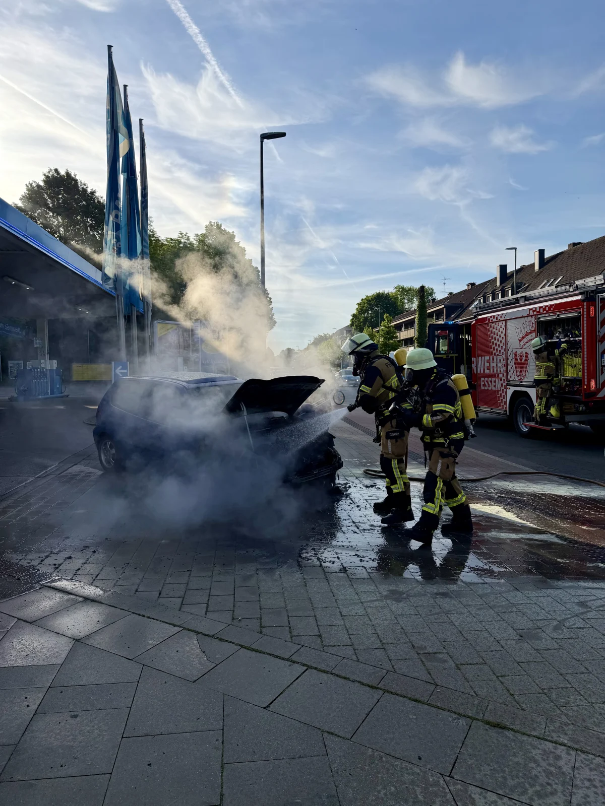 Zwei Feuerwehrleute in Schutzkleidung löschen ein brennendes Auto, aus dem Rauch aufsteigt. Ein Feuerwehrauto steht im Hintergrund.