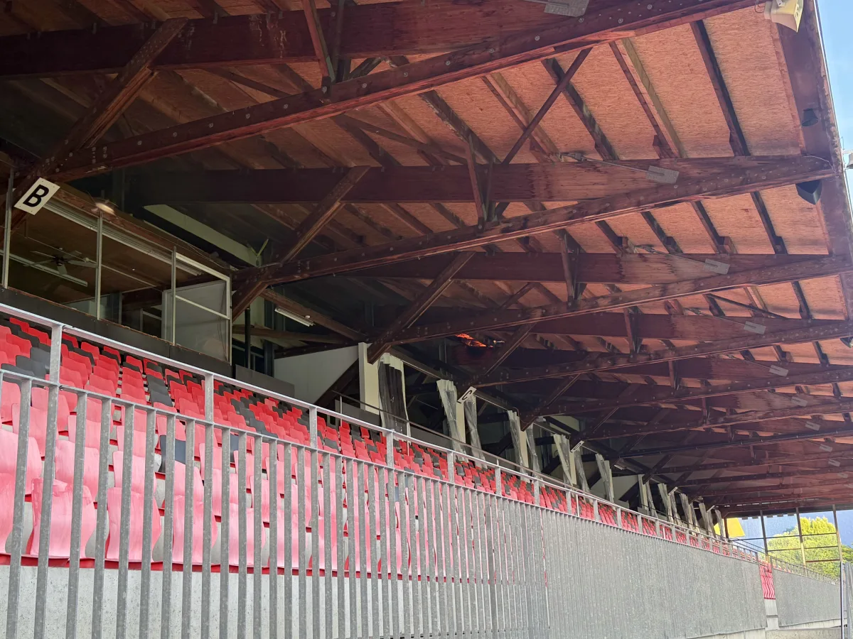 Eine leere Tribüne mit Holzdach und roten Sitzen in einem Stadion. Im Hintergrund sind Teile der Gebäude der Umgebung sichtbar.