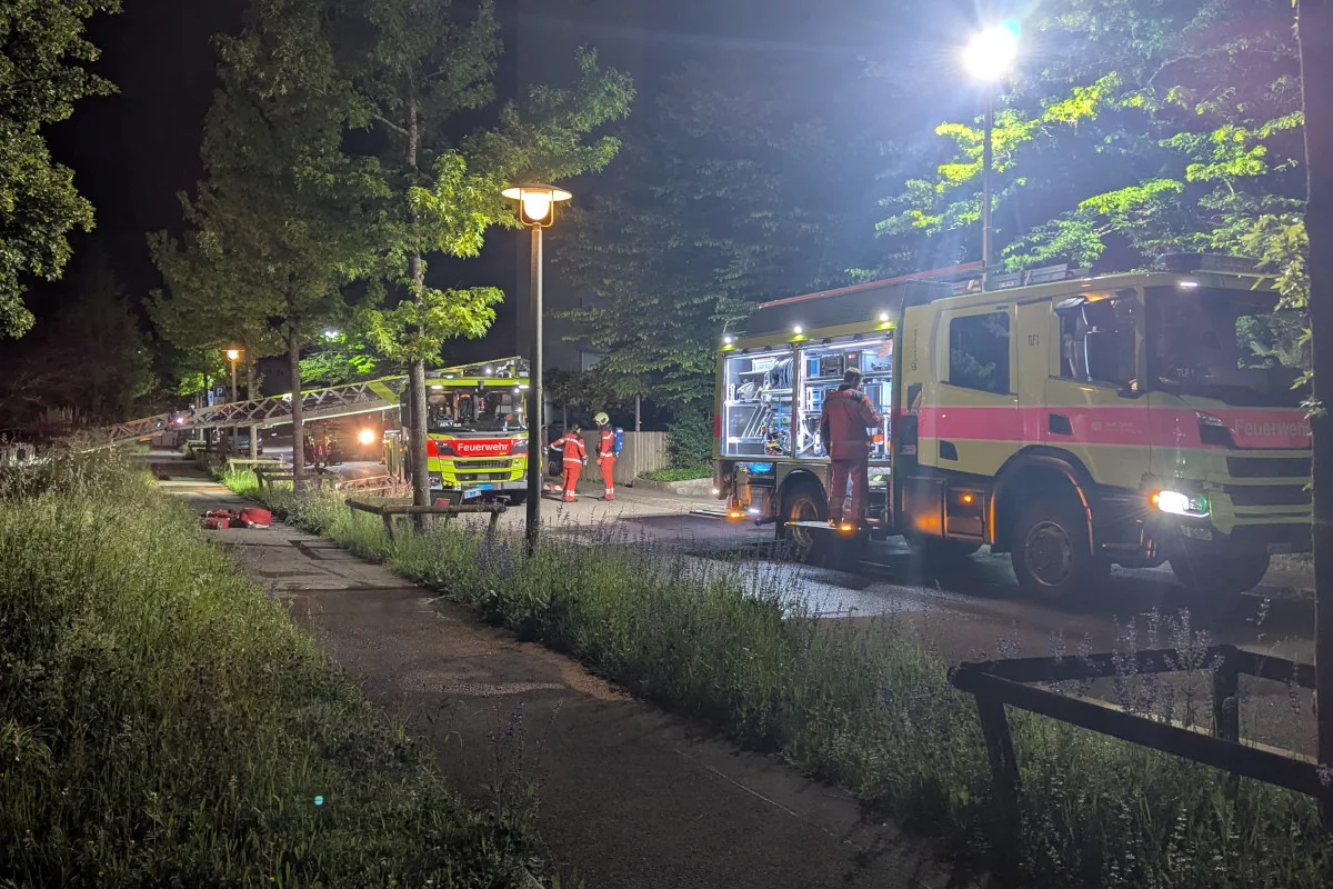 Zwei Feuerwehrfahrzeuge stehen nachts auf einem Gehweg, umgeben von Bäumen und beleuchtet durch Straßenlampen. Einsatzkräfte in roten Uniformen sind bei der Arbeit zu sehen.
