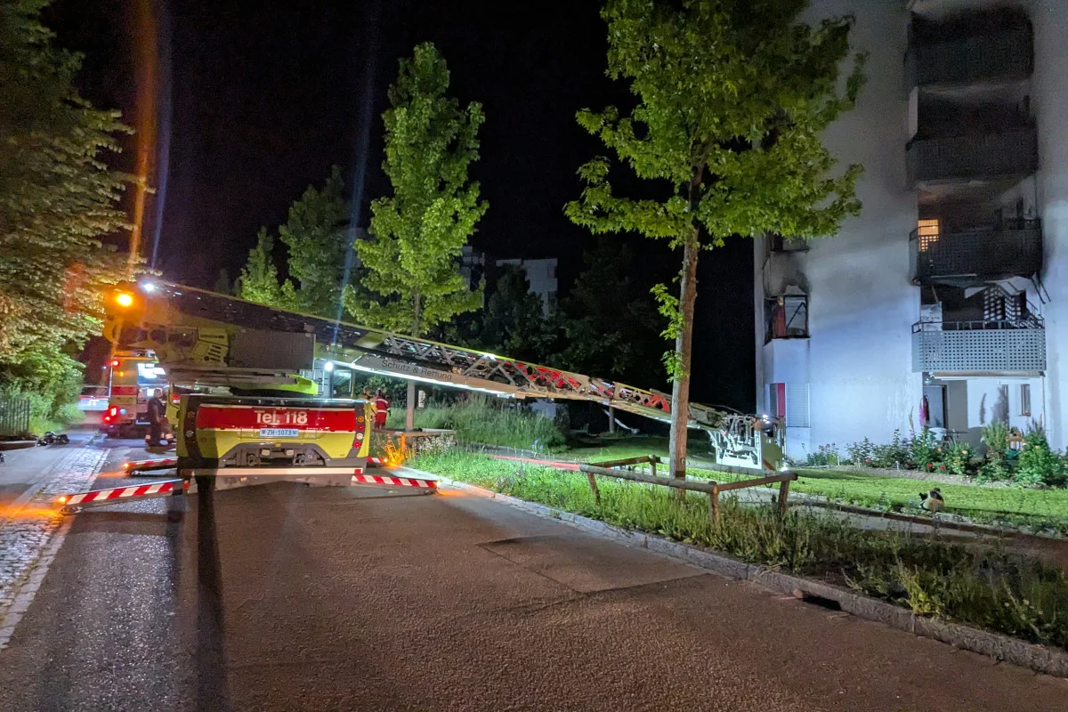 Ein Feuerwehrfahrzeug mit ausgefahrener Leiter steht nachts vor einem Wohnhaus, dessen Fassade Brandspuren zeigt.