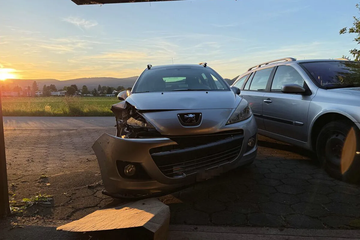 Ein Auto mit beschädigter Front steht auf einer Einfahrt; der Sonnenuntergang ist im Hintergrund sichtbar. Ein weiteres Auto ist rechts daneben geparkt.