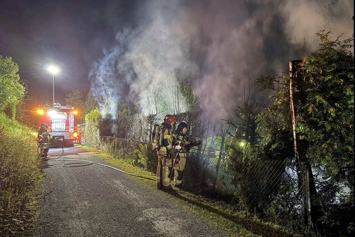 Bei Nacht sind Feuerwehrleute zu sehen, die an einem Zaun einen Brand löschen. Ein Löschfahrzeug beleuchtet die Szene.