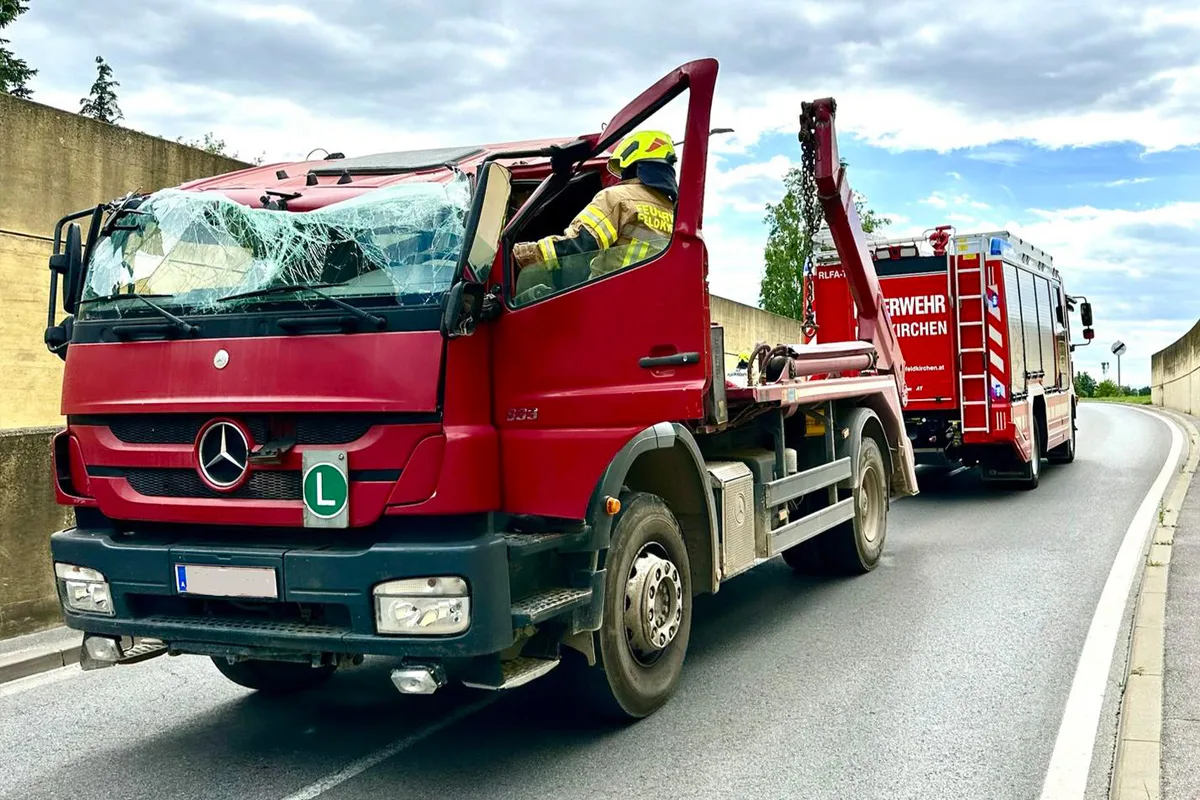 Ein roter Mercedes-LKW mit zerbrochener Windschutzscheibe steht auf einer Straße. Ein Feuerwehrmann ist in der Kabine, dahinter ein Feuerwehrfahrzeug.
