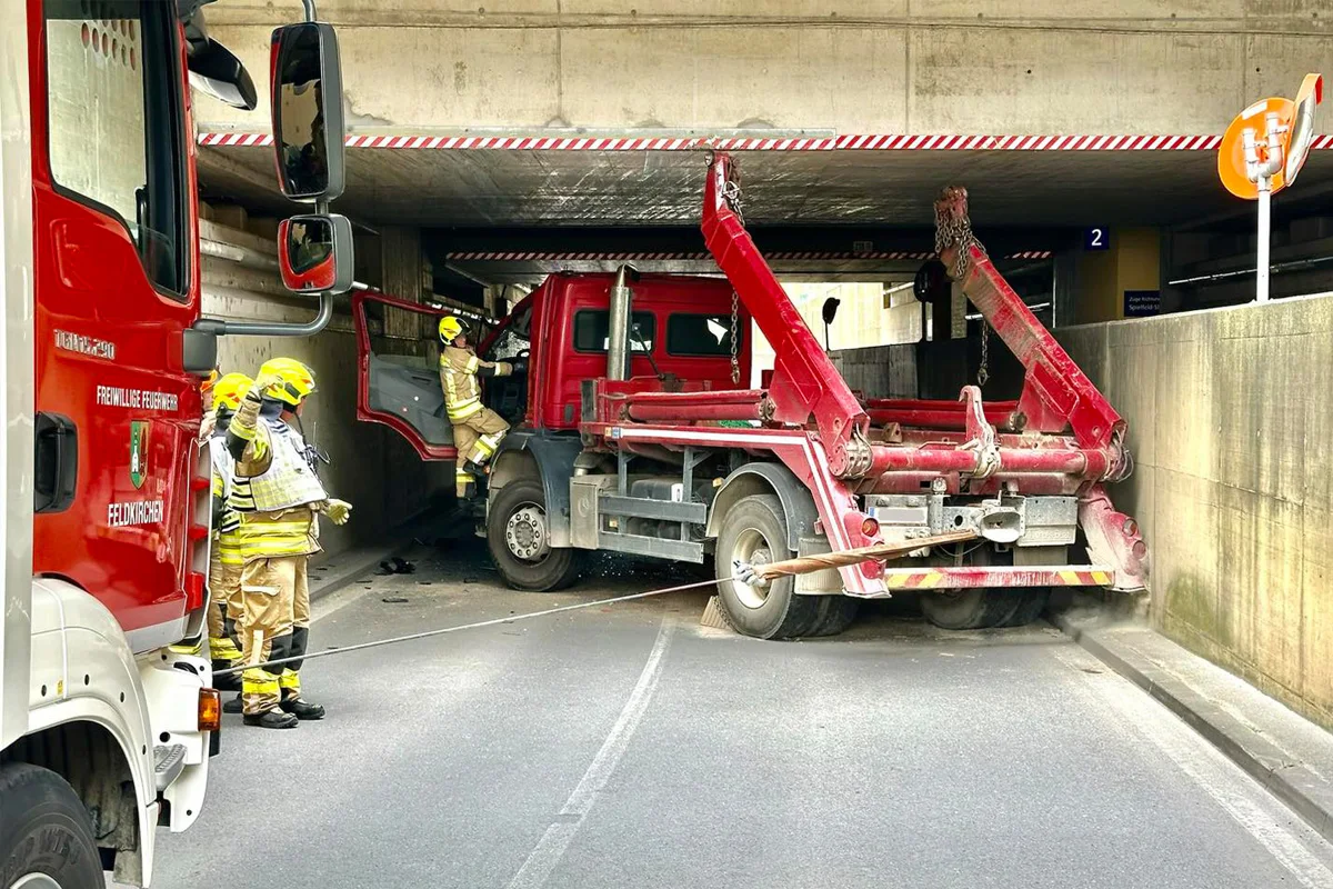 Ein roter Lastwagen hat sich unter einer Brücke festgefahren, während Feuerwehrleute in gelber Schutzkleidung versuchen zu helfen.