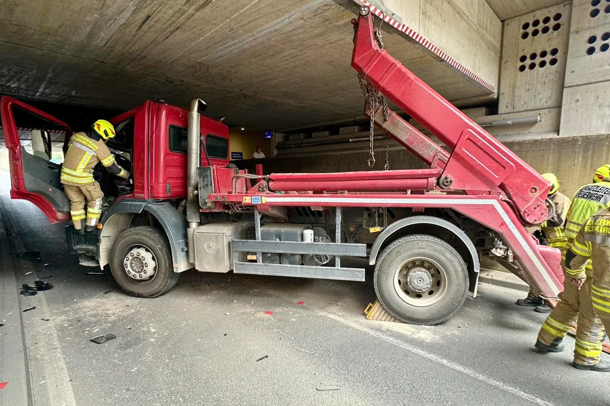 Ein roter Lastwagen ist unter einer Brücke eingeklemmt, während zwei Feuerwehrleute in neongelber Schutzkleidung Hilfe leisten. Der Innenraum des Tunnels ist dunkel und die Betonbrücke zeigt keine sichtbaren Schäden.