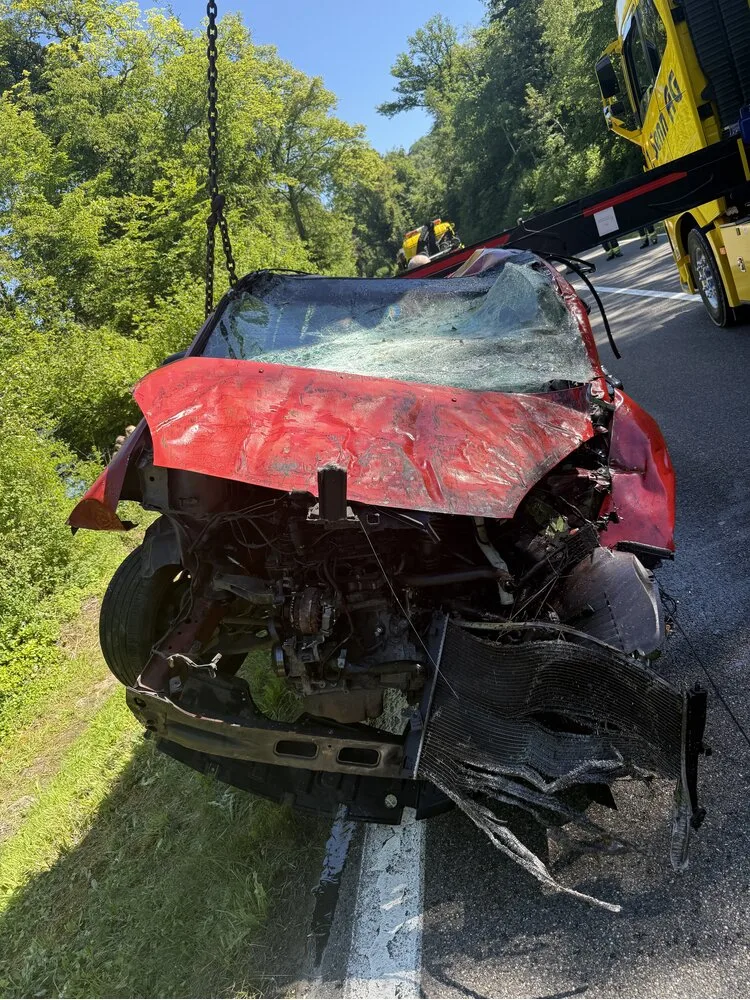 Ein rotes Auto mit starkem Frontschaden wird auf einer Straße von einem Abschleppfahrzeug angehoben. Es ist Tageslicht und es gibt viel Grün an der Straßenseite.