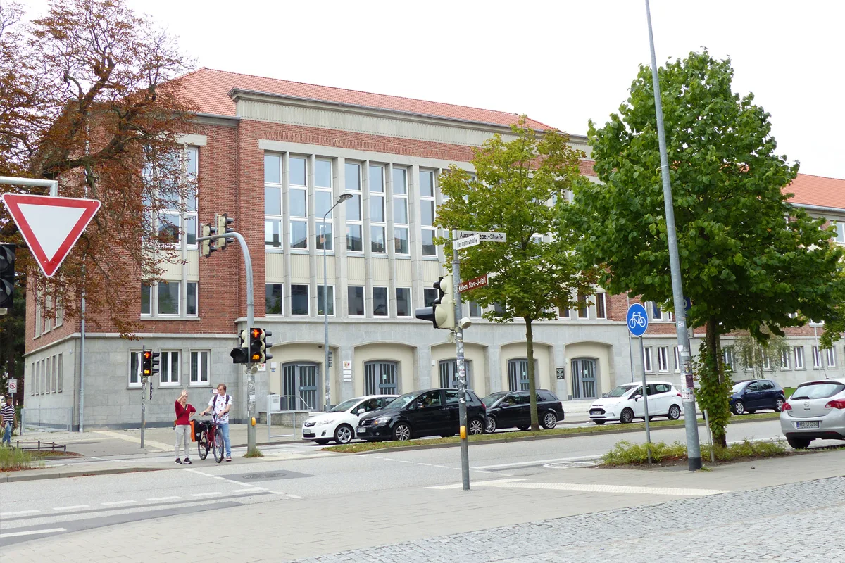 Ein großes, rotes Backsteingebäude steht neben einer Kreuzung, an der mehrere Autos parken und zwei Fußgänger mit einem Fahrrad bei einer roten Ampel stehen.