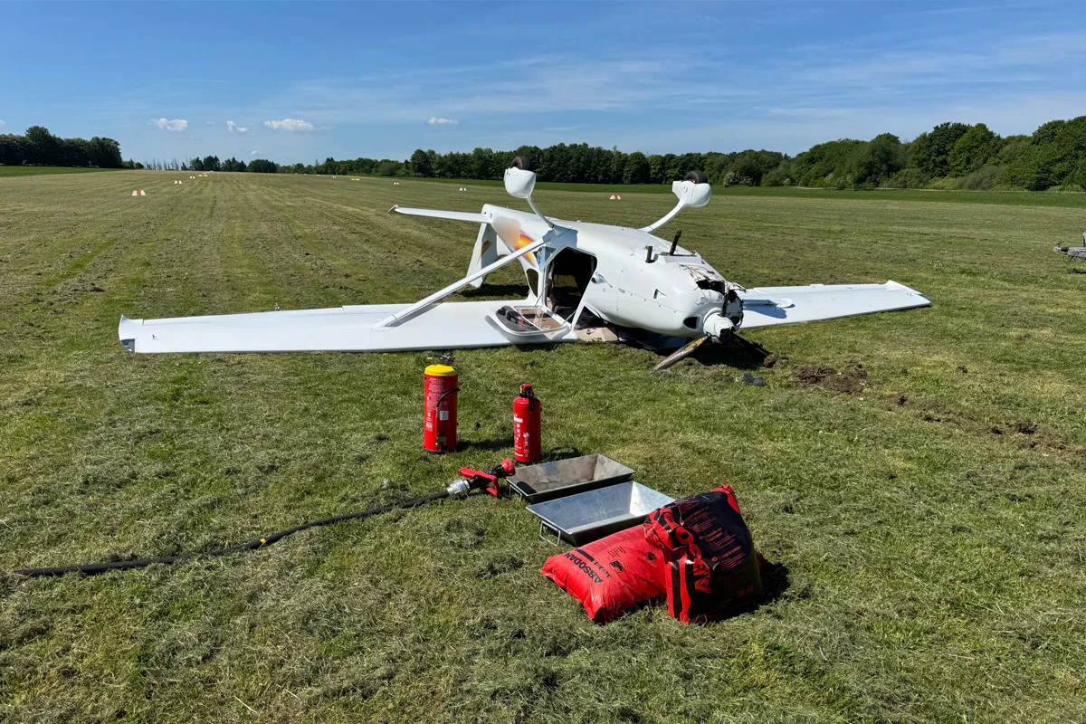 Ein weißes Kleinflugzeug liegt abgestürzt auf einem offenen Feld. Rundherum liegen zwei Feuerlöscher und weiteres Löschmaterial.