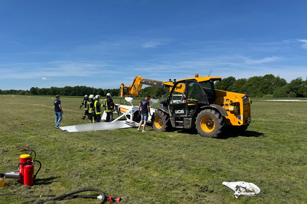 Mehrere Feuerwehrleute stehen auf einer Wiese um ein umgekipptes Flugzeugwrack. Ein gelber Bagger ist in der Nähe.