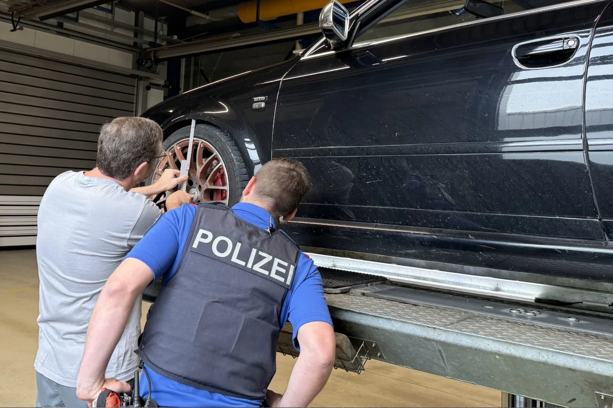 Ein Polizist und ein Zivilist untersuchen gemeinsam das Rad eines schwarzen Autos, das auf einer Hebebühne in einer Werkstatt steht. Der Polizist trägt eine Weste mit 'Polizei'-Aufschrift.