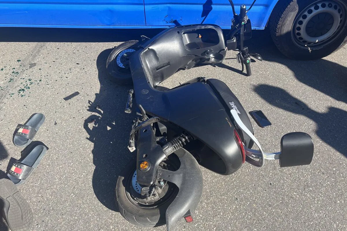 Ein schwarzer E-Scooter liegt beschädigt auf der Straße neben einem blauen Fahrzeug. Auf dem Boden befinden sich Scherben und ein Paar Sandalen.
