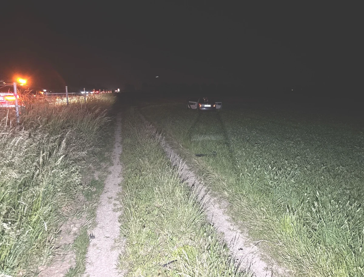 Ein Auto ist bei Nacht auf einem Feldweg neben einer Landstraße geparkt. Die Straße ist beleuchtet, während das Auto im Dunkeln steht.