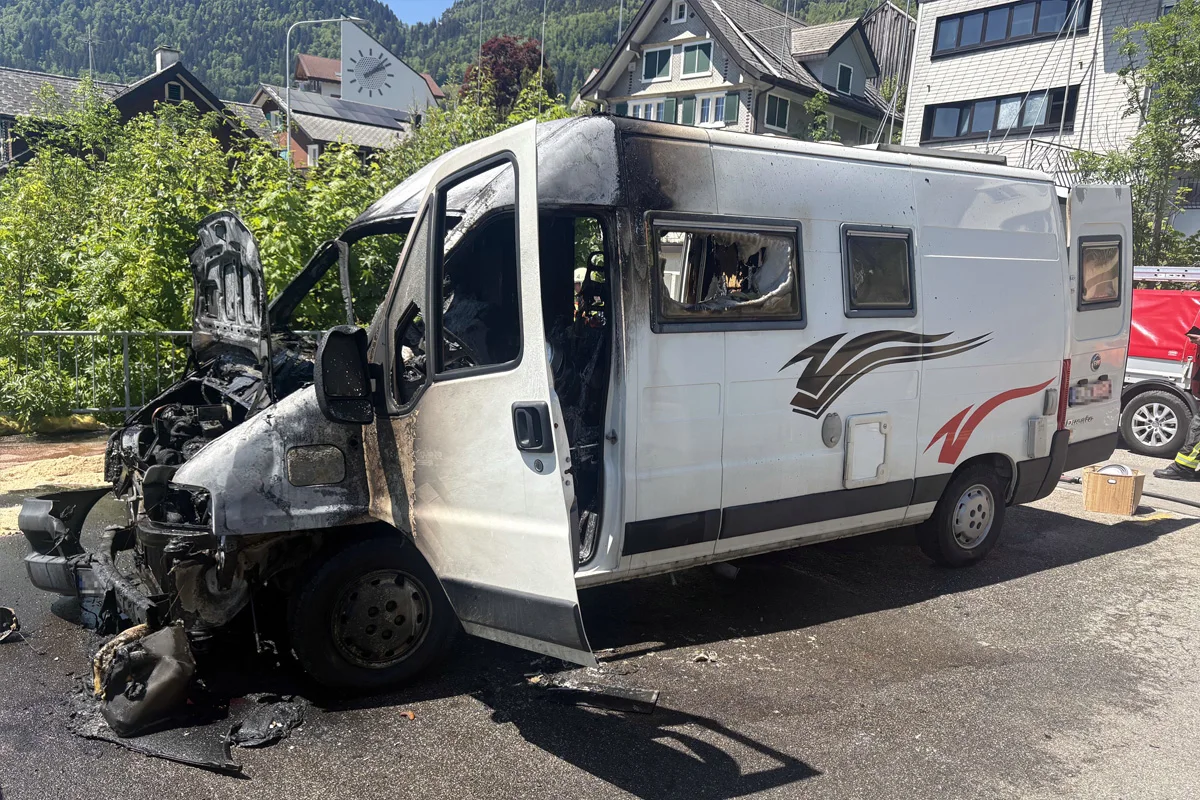 Ein weißer Kleintransporter, dessen Vorderteil und Innenraum stark ausgebrannt sind, steht auf einer Straße in einem Wohngebiet. Im Hintergrund sind Bäume und Wohnhäuser zu sehen.