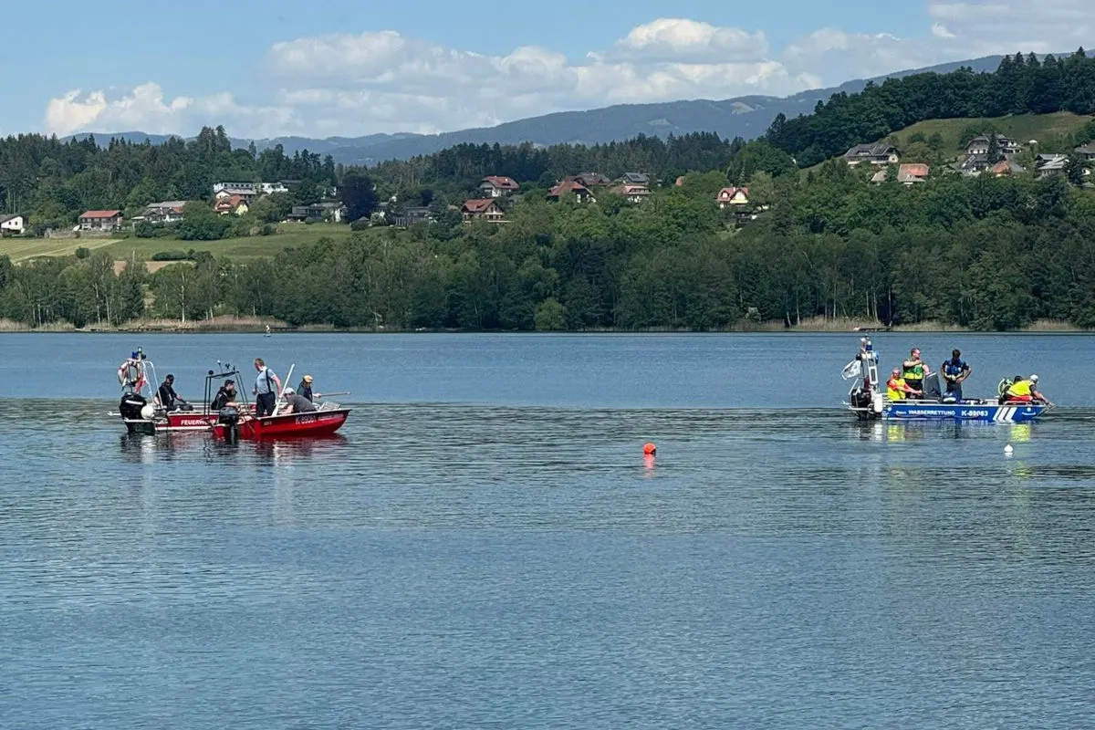 Zwei Boote mit Rettungskräften stehen auf einem See, das eine Boot ist rot und markiert, während das andere blau ist. Im Hintergrund sind Hügel und Häuser sichtbar, es ist klares Wetter.