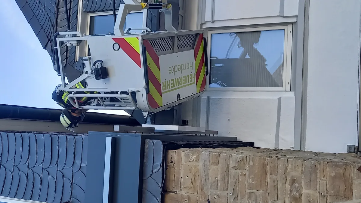 Ein Feuerwehrmann in einem erhöhten Rettungskorb ist auf Höhe eines Gebäudefensters zu sehen. Das Fahrzeug hat deutliche Markierungen der Feuerwehr.