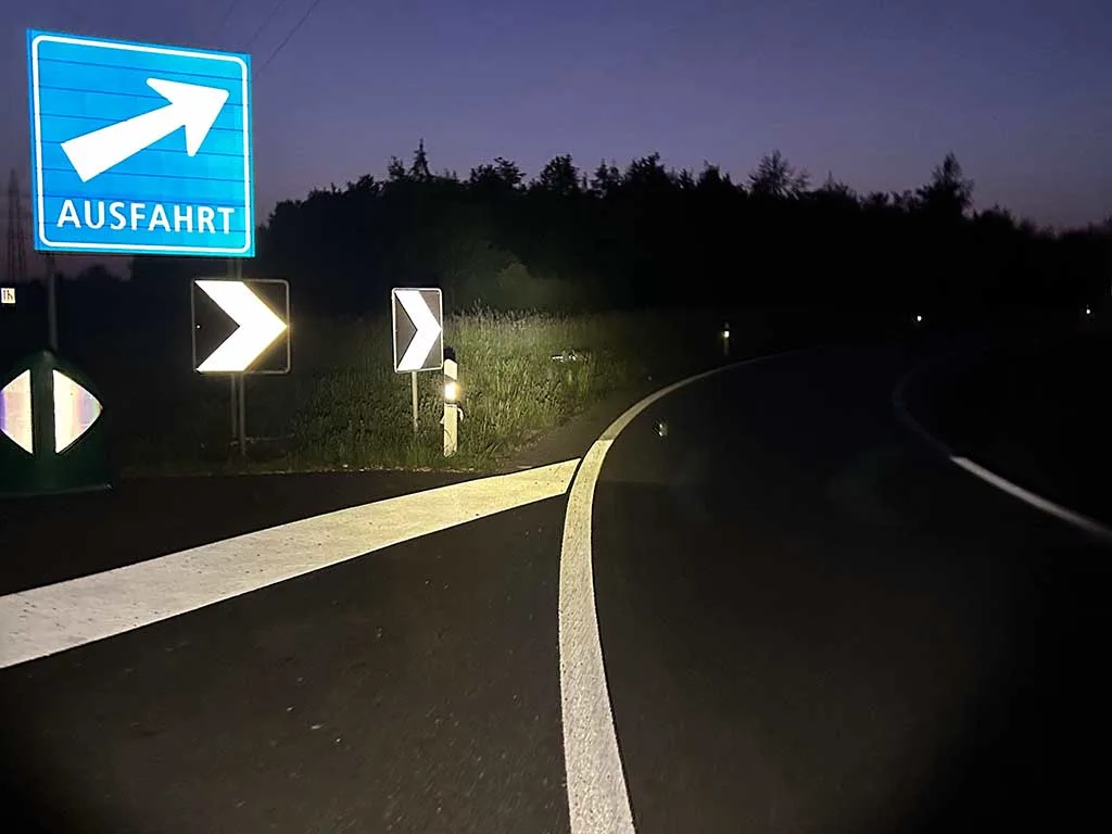 Ein blaues Ausfahrtsschild zeigt nach rechts auf einer dunklen Autobahnausfahrt. Die Straße ist durch Fahrzeuglichter beleuchtet.