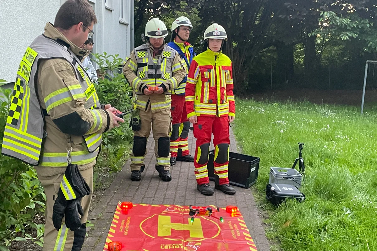 Drei Feuerwehrleute stehen auf einem Gehweg neben einem roten Drohnen-Landeplatz. Sie tragen Schutzkleidung, Helme und halten Fernsteuerungen.
