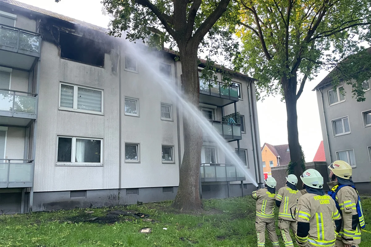Mehrere Feuerwehrleute in Schutzkleidung löschen mit einem Wasserstrahl ein brennendes Mehrfamilienhaus. Die Fassade ist beschädigt.