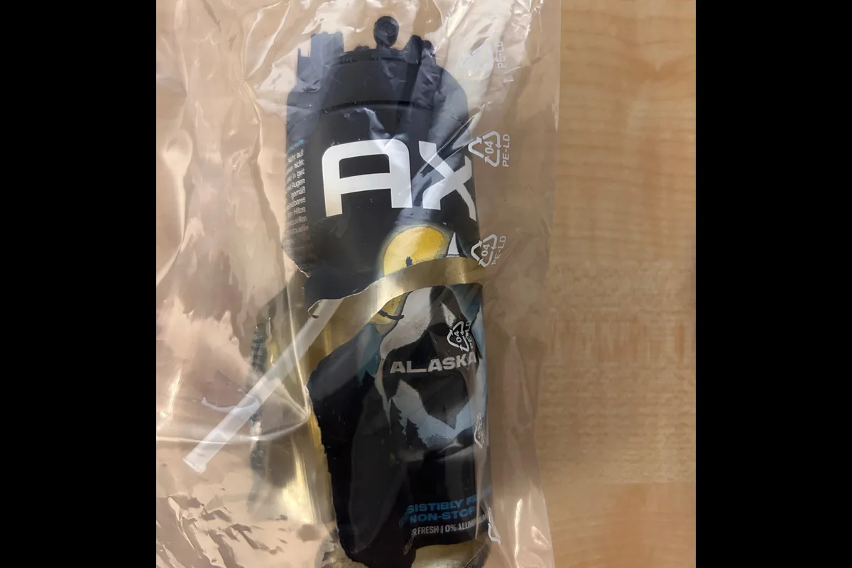 Eine schwarze Dose mit der Aufschrift 'Axe Alaska' steckt in einer transparenten Kunststoffverpackung. Der Hintergrund ist aus Holz.