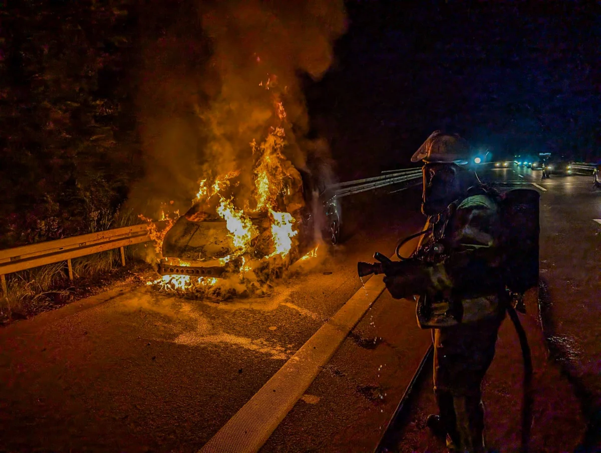 Auf einer nächtlichen Autobahn steht ein Auto in Flammen. Ein Feuerwehrmann in Schutzkleidung versucht, das Feuer zu löschen.