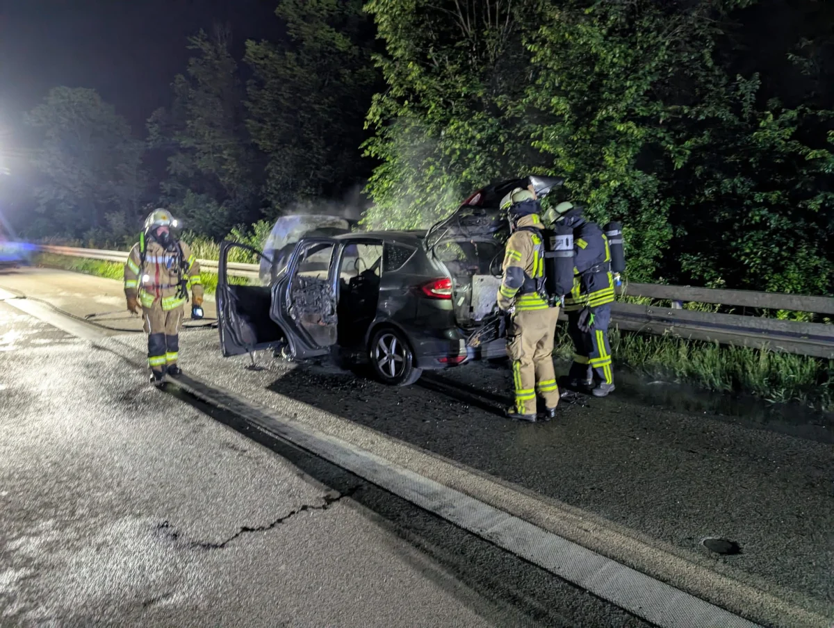 Drei Feuerwehrleute stehen neben einem ausgebrannten Auto auf einer Autobahn bei Nacht; im Hintergrund Bäume erkennbar.