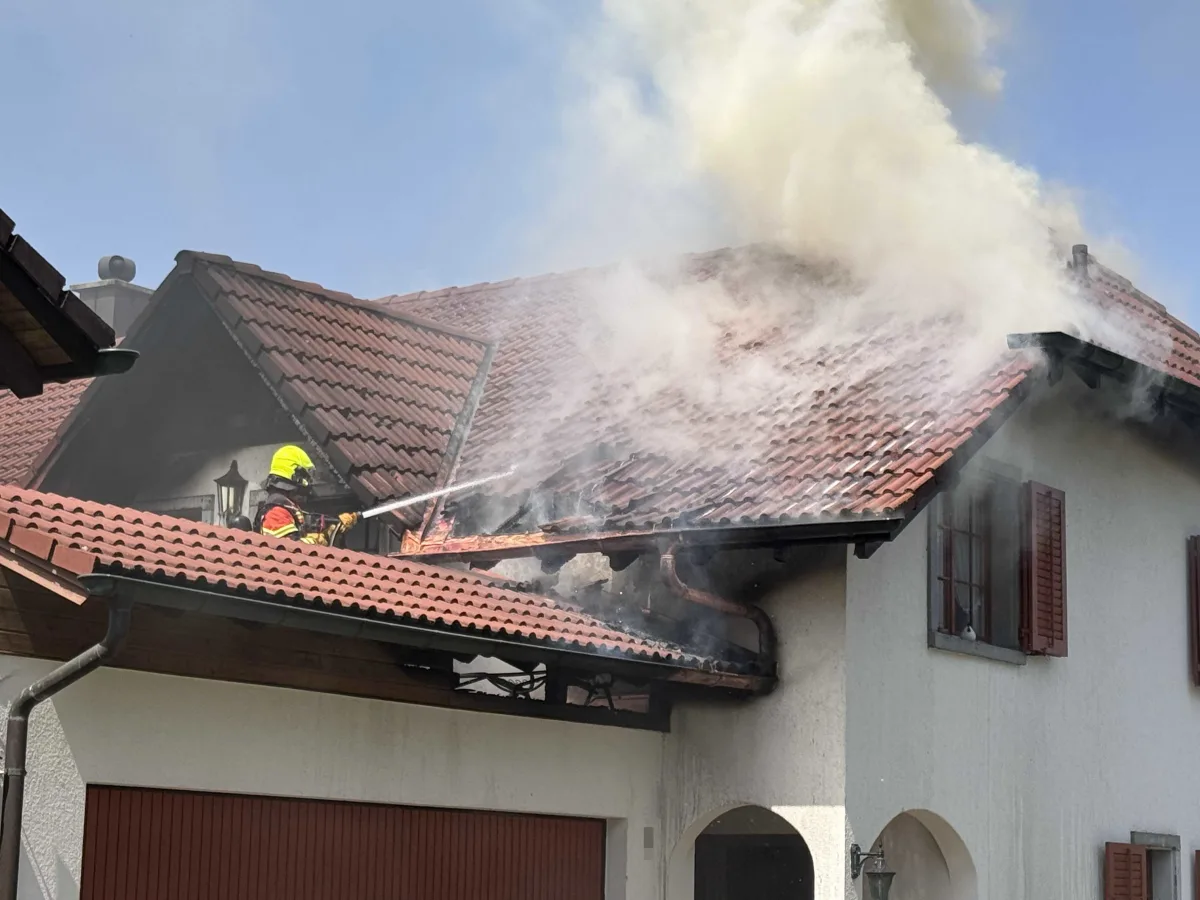 Ein Feuerwehrmann in Schutzkleidung spritzt Wasser auf einen rauchenden Dachstuhl eines Wohnhauses. Dichter Rauch steigt auf.