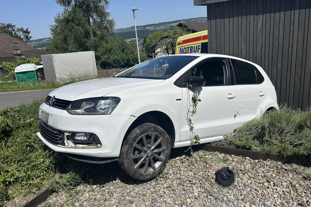 Ein weißer VW ist in eine Hecke gefahren und steht schief. Im Hintergrund ist ein Notrufwagen zu sehen. Die Unfallstelle befindet sich in einem ländlichen Gebiet.