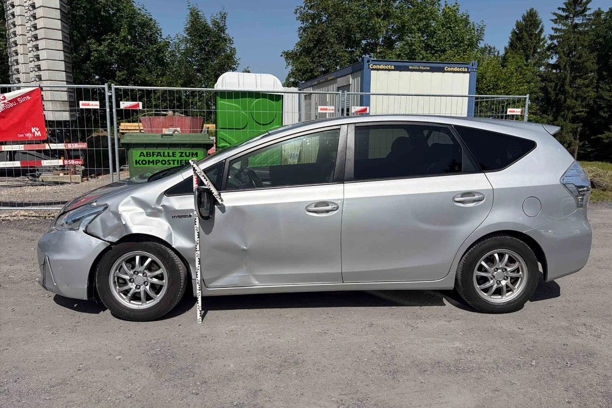 Das Bild zeigt ein silbernes Auto mit einer stark eingedrückten Fahrertür, das auf einem Parkplatz vor einem Bauzaun steht. Im Hintergrund sind Bäume und Baucontainer zu sehen.