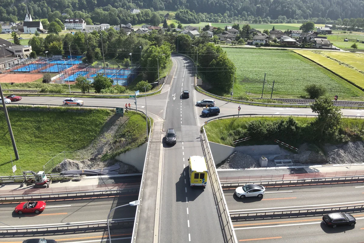 Mehrere Fahrzeuge bewegen sich über eine Überführung, die eine Autobahn kreuzt, umgeben von grünen Feldern und Gebäuden.