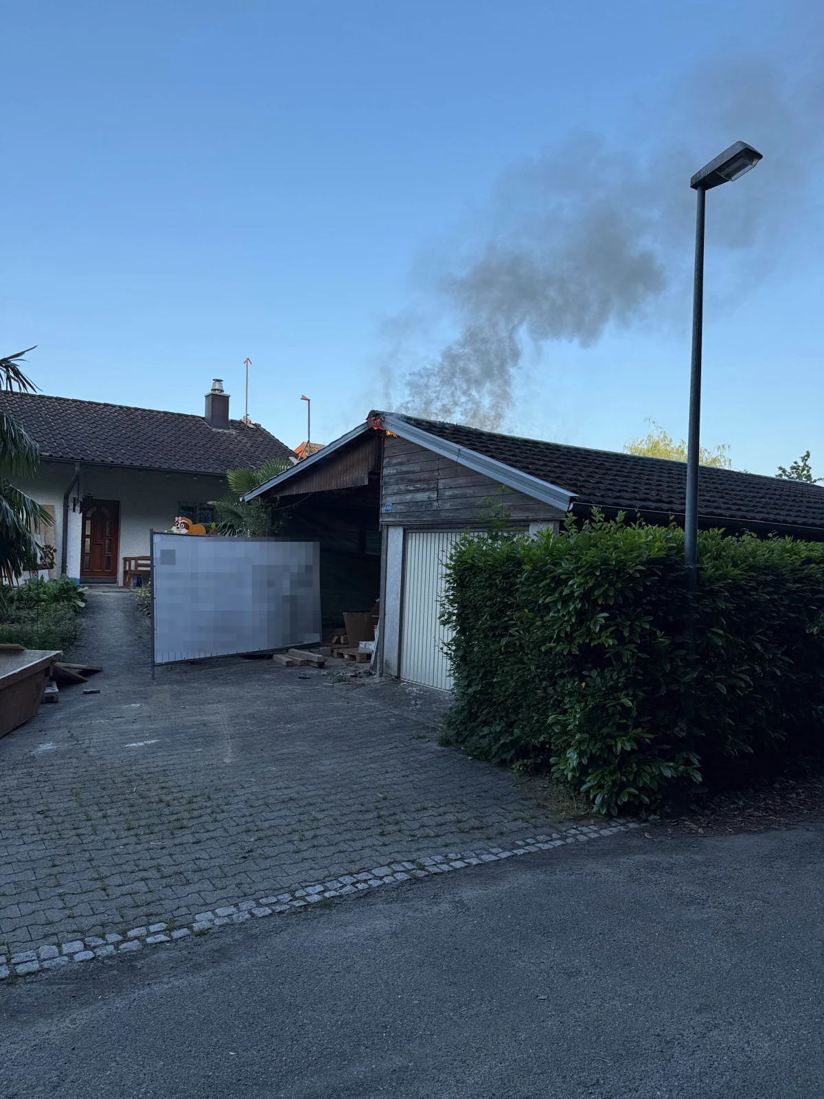 Eine Garage in einem Wohngebiet zeigt Rauchschwaden, die vom Dach aufsteigen, während daneben ein Haus steht.