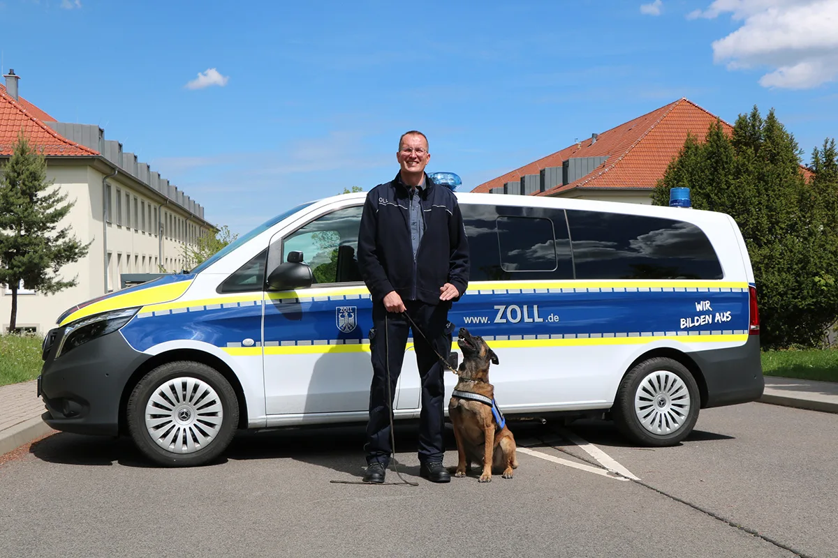 Ein Zollbeamter steht auf einer Straße vor einem weißen und blauen Zollfahrzeug. Neben ihm sitzt ein Hund.