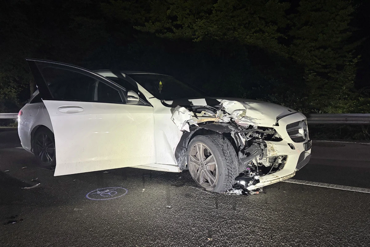 Ein weißer Mercedes mit beschädigter Front steht mit geöffneter Fahrertür auf einer dunklen Straße. Das Auto zeigt deutliche Unfallspuren.