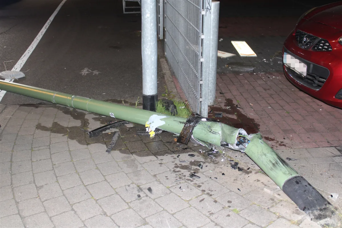 Eine grüne Laterne ist umgeknickt und liegt auf dem Boden neben einem roten Auto am Straßenrand. Teile der Laterne sind verbogen und beschädigt.