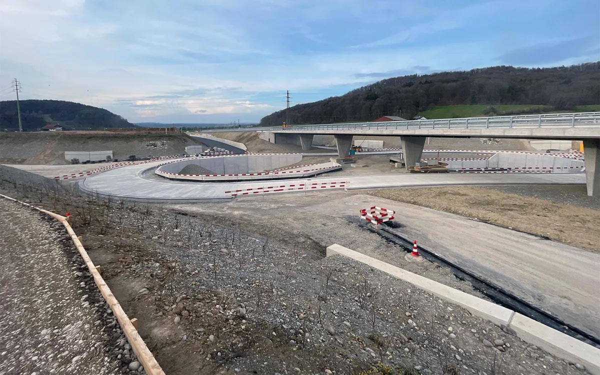 Eine unvollendete Autobahnunterführung mit roten und weißen Absperrungen ist sichtbar. Im Hintergrund sind Hügel und Stromleitungen zu erkennen.
