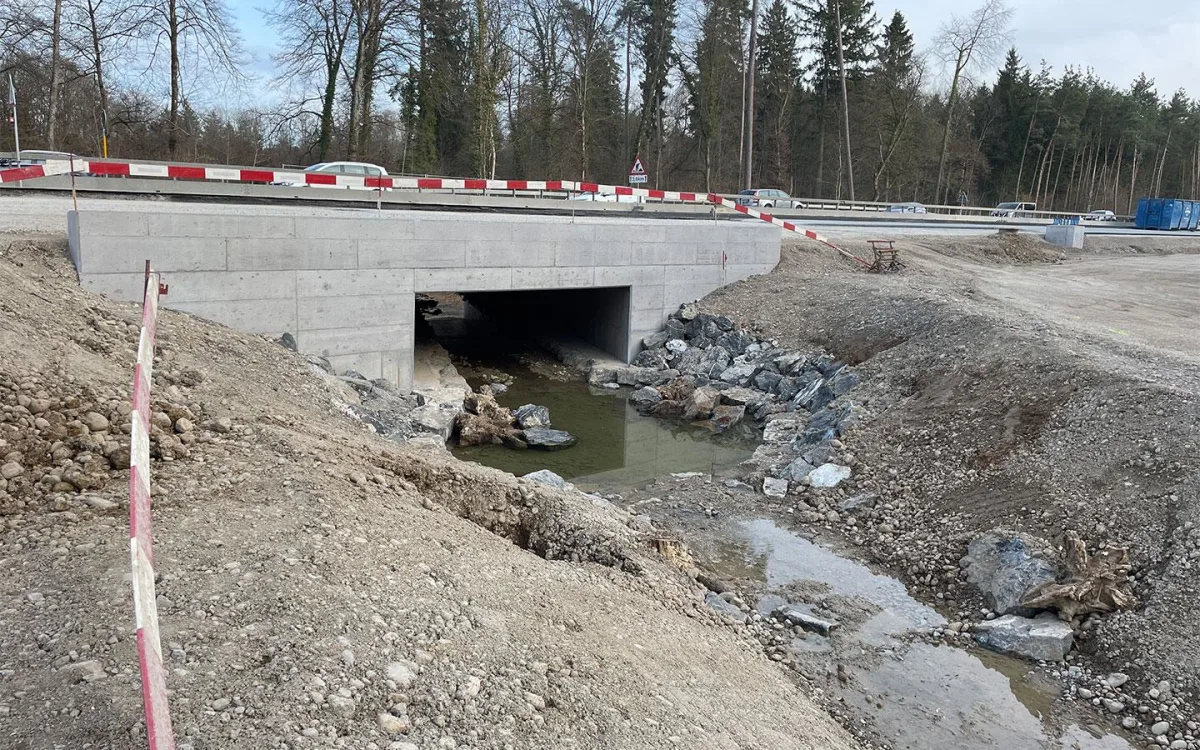 Eine Betonbrücke führt über einen kleinen, fließenden Bach, umgeben von Erdarbeiten und abgesichert durch provisorische Barrieren.