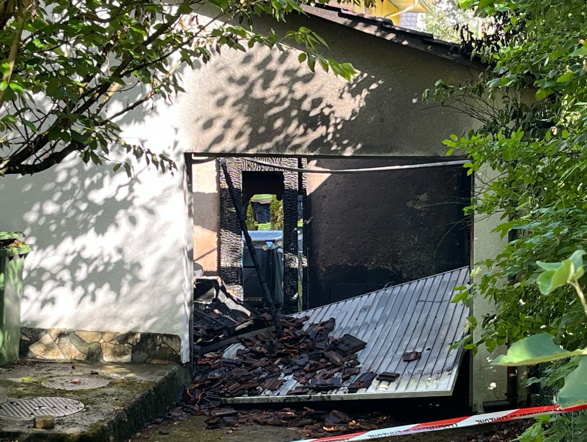 Ein zerstörter Eingang zeigt ein verkohltes Loch in der Wand, umgeben von umgefallenem Schutt und einem umgestürzten Tor. Bäume rahmen die Szene ein.