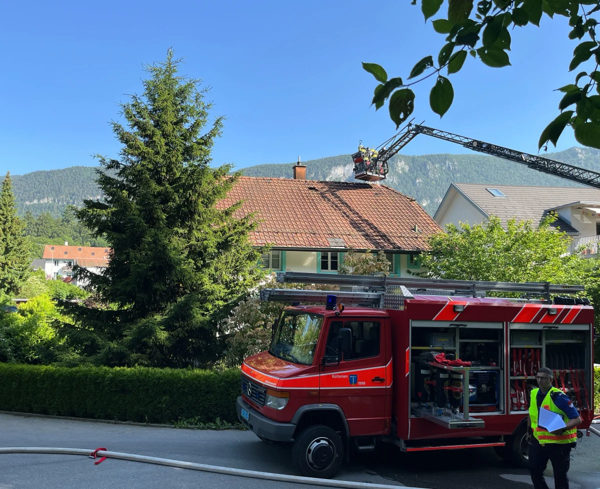 Ein rotes Feuerwehrfahrzeug steht vor einem Haus mit Einsatzkräften auf dem Dach. Im Hintergrund sind Bäume und Berge sichtbar.