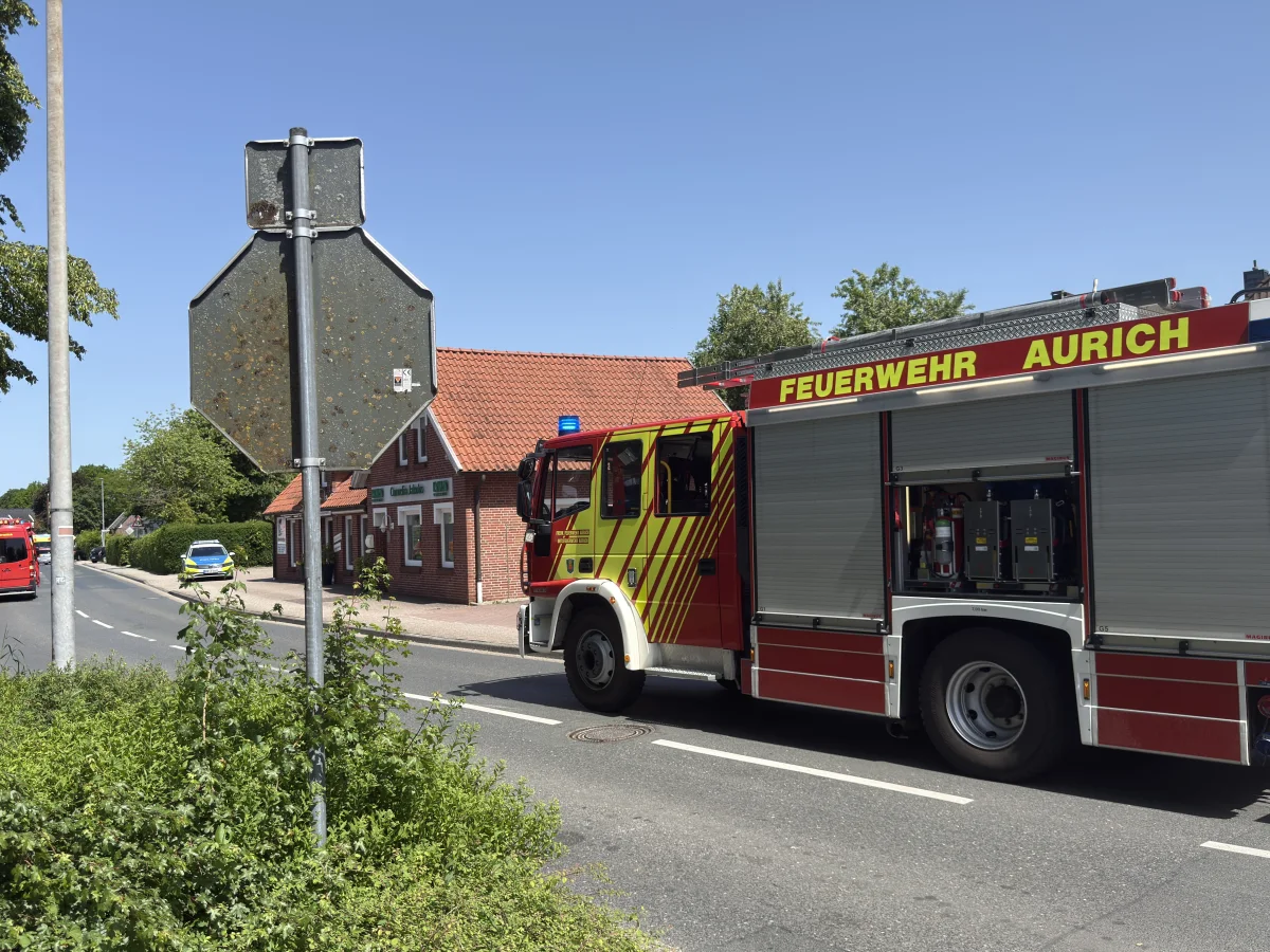 Ein Feuerwehrfahrzeug mit der Aufschrift 'FEUERWEHR AURICH' steht auf einer Straße. Daneben ist ein Backsteingebäude zu sehen, und ein weiteres Einsatzfahrzeug ist im Hintergrund erkennbar. Es ist sonnig.