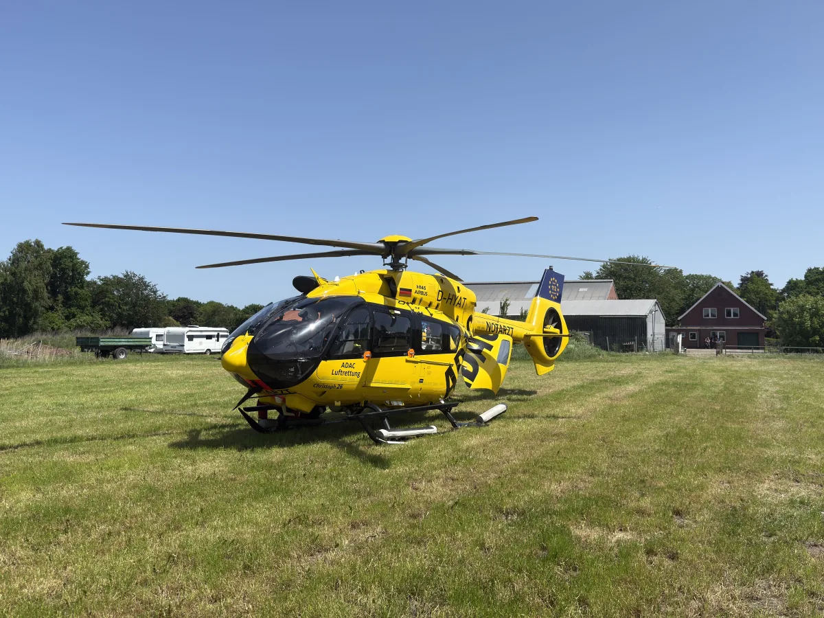 Ein gelber ADAC-Rettungshubschrauber steht auf einer Wiese, im Hintergrund ist ein Wohnhaus und einige Bäume zu sehen. Der Himmel ist klar und strahlend blau.