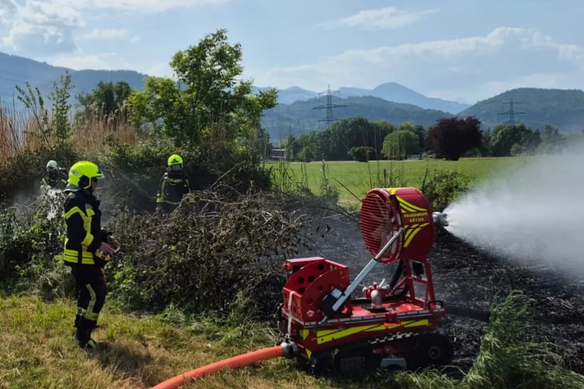 Feuerwehrleute nutzen ein motorisiertes Löschgerät, um einen Brand auf einer Wiese unter Kontrolle zu bringen. Im Hintergrund sind Berge sichtbar, und die Szene findet bei klarem Wetter statt.