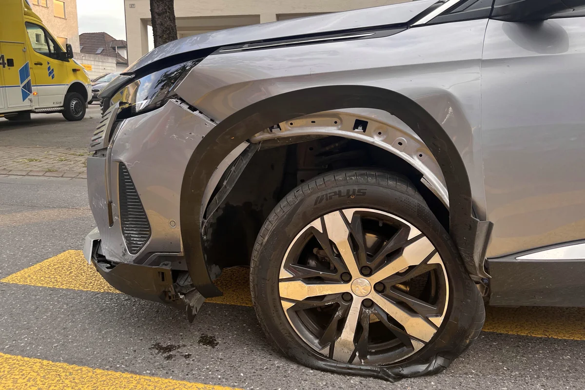Ein silbernes Auto zeigt einen abgeflachten und beschädigten Vorderreifen. Im Hintergrund ist ein Rettungsfahrzeug sichtbar.