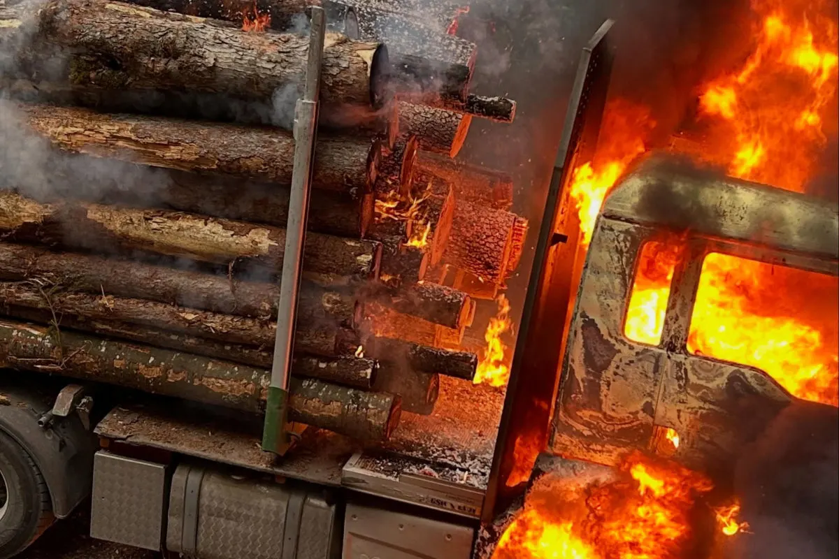 Das Bild zeigt einen Holzlaster, dessen Fahrerhaus in Flammen steht. Dichte Rauchwolken steigen auf, während die Baumstämme ebenfalls Feuer fangen.