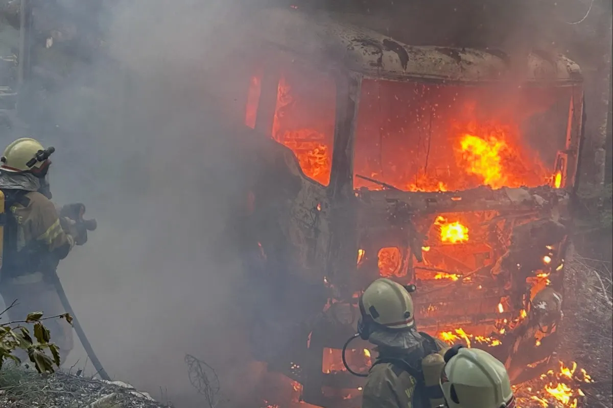 Mehrere Feuerwehrleute kämpfen gegen einen intensiven Fahrzeugbrand im Wald, umgeben von dicken Rauchschwaden und Flammen.