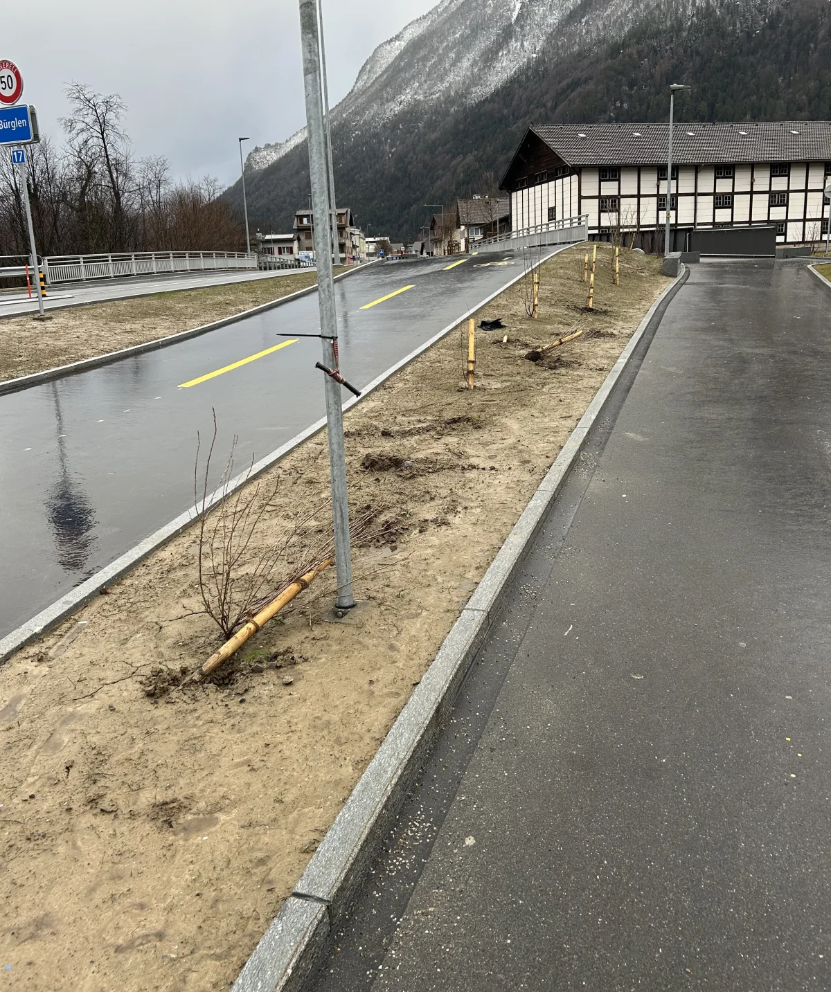 Ein Verkehrsschild auf einer leeren, nassen Straße in einer ländlichen Umgebung mit Bergen im Hintergrund. Der Boden ist matschig, und einige junge Bäume sind an Pfosten gebunden.