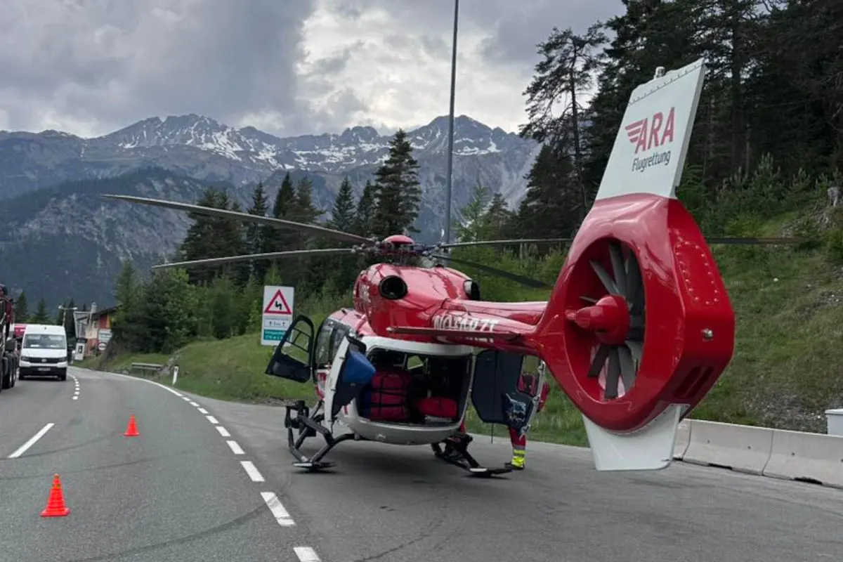 Ein roter Rettungshubschrauber steht auf einer Bergstraße mit geöffneter Tür. Der Hintergrund zeigt bewaldete Berge und Wolken am Himmel.
