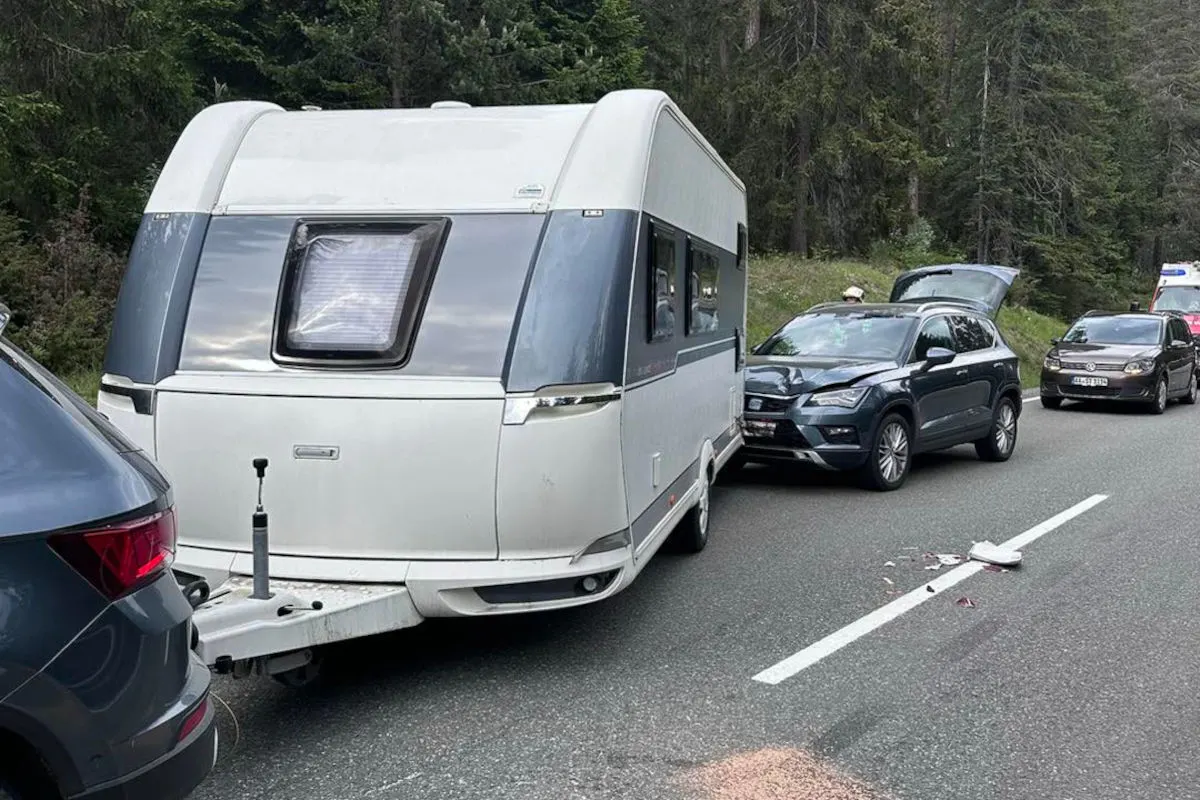 Ein Wohnwagen ist in einen Unfall verwickelt, bei dem zwei Autos auf einer Landstraße auffahren. Es gibt sichtbare Schäden an den Fahrzeugen und Trümmer auf der Straße.