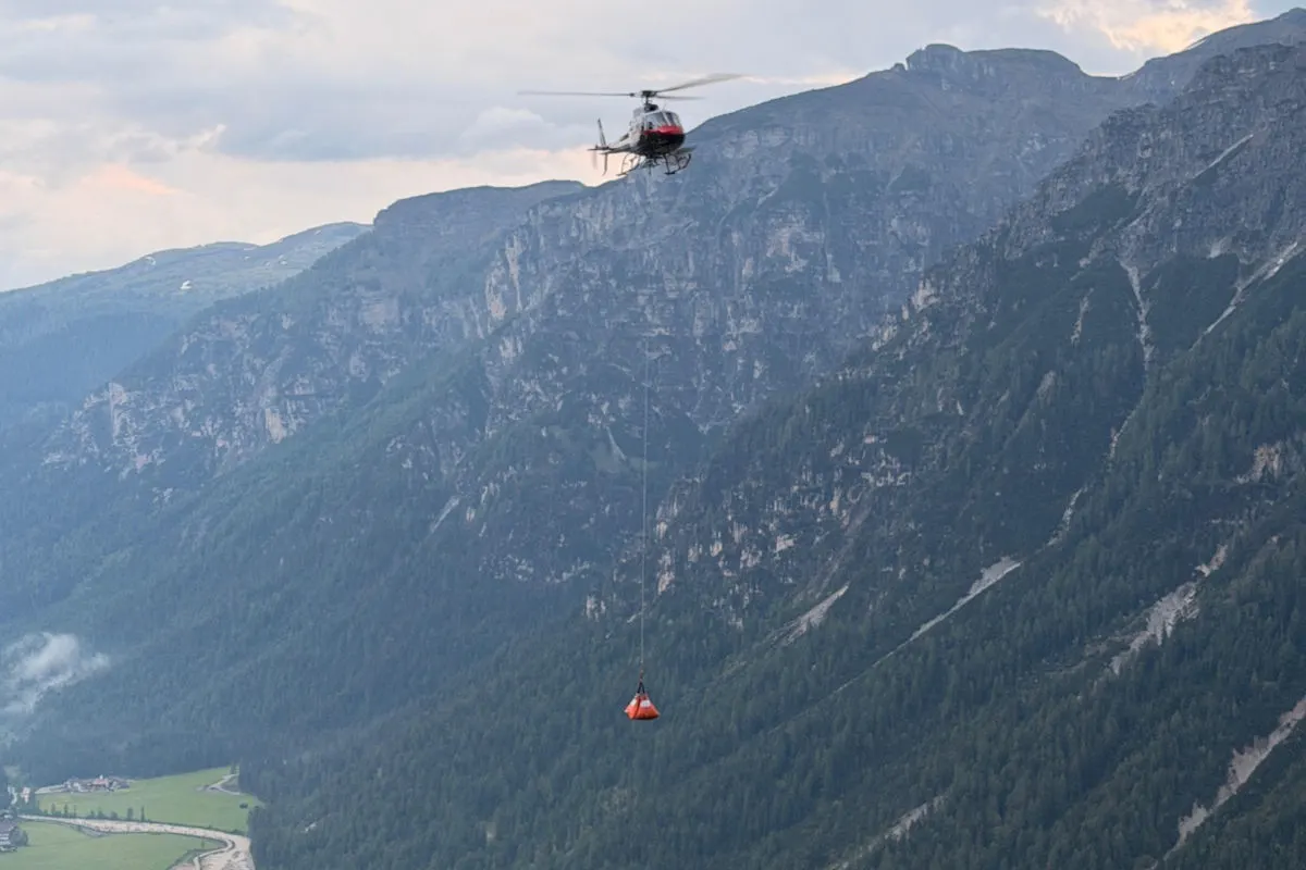 Ein Hubschrauber schwebt mit einem roten Löschbehälter über einer weitläufigen Berglandschaft. Der Hintergrund zeigt hohe, bewaldete Berge.
