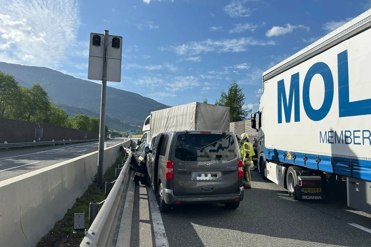 Ein grauer Kleintransporter ist zwischen einer Leitplanke und einem weißen Lkw auf einer Autobahn eingeklemmt. Im Hintergrund sind einige Berge und ein blauer Himmel zu sehen.